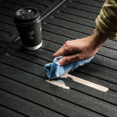 Close-up of a hand wiping a beige liquid spill on a dark ribbed surface with a blue microfiber cloth; a takeaway coffee cup sits nearby.