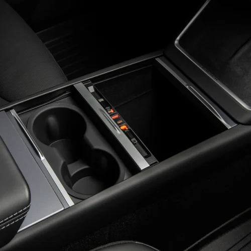 Close-up of a car center console with two cup holders and a sliding storage tray with orange-lit charging ports inside a dark interior