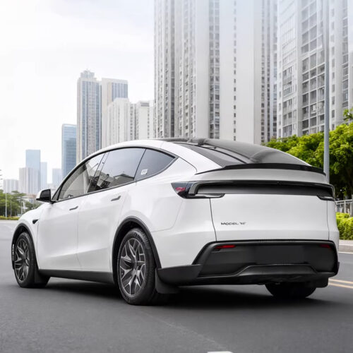 Rear three-quarter view of a white Tesla Model Y electric SUV on a city street with tall modern buildings in the background.