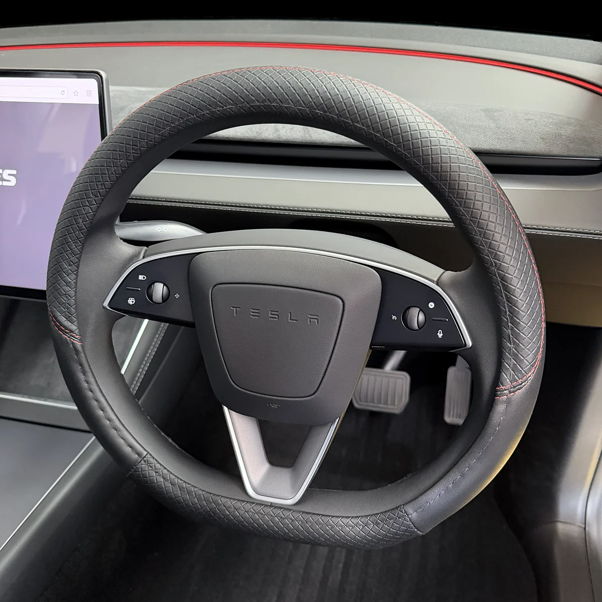 Tesla steering wheel with textured grip and red stitching, multifunction controls on both sides, and a centered TESLA badge; pedals visible below.