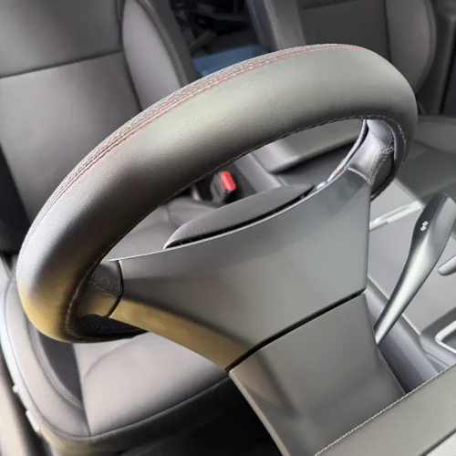 Close-up of a car steering wheel with red contrast stitching on black leather, showing grip and spokes inside the cabin