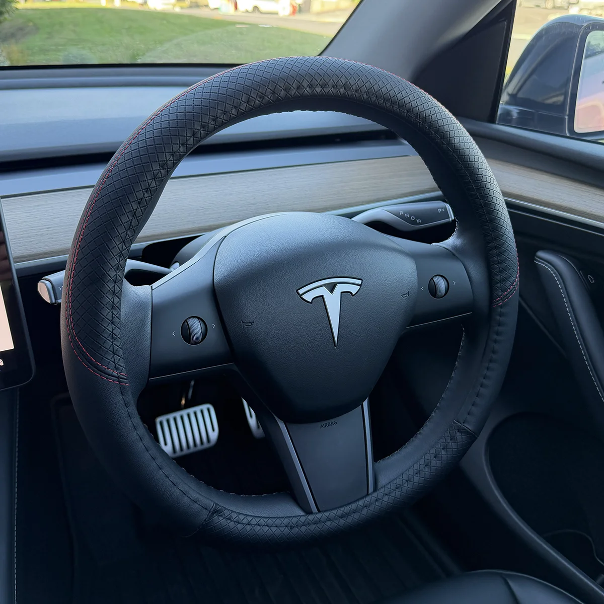 Tesla steering wheel with textured black cover and red stitching, dashboard visible behind in a modern car interior