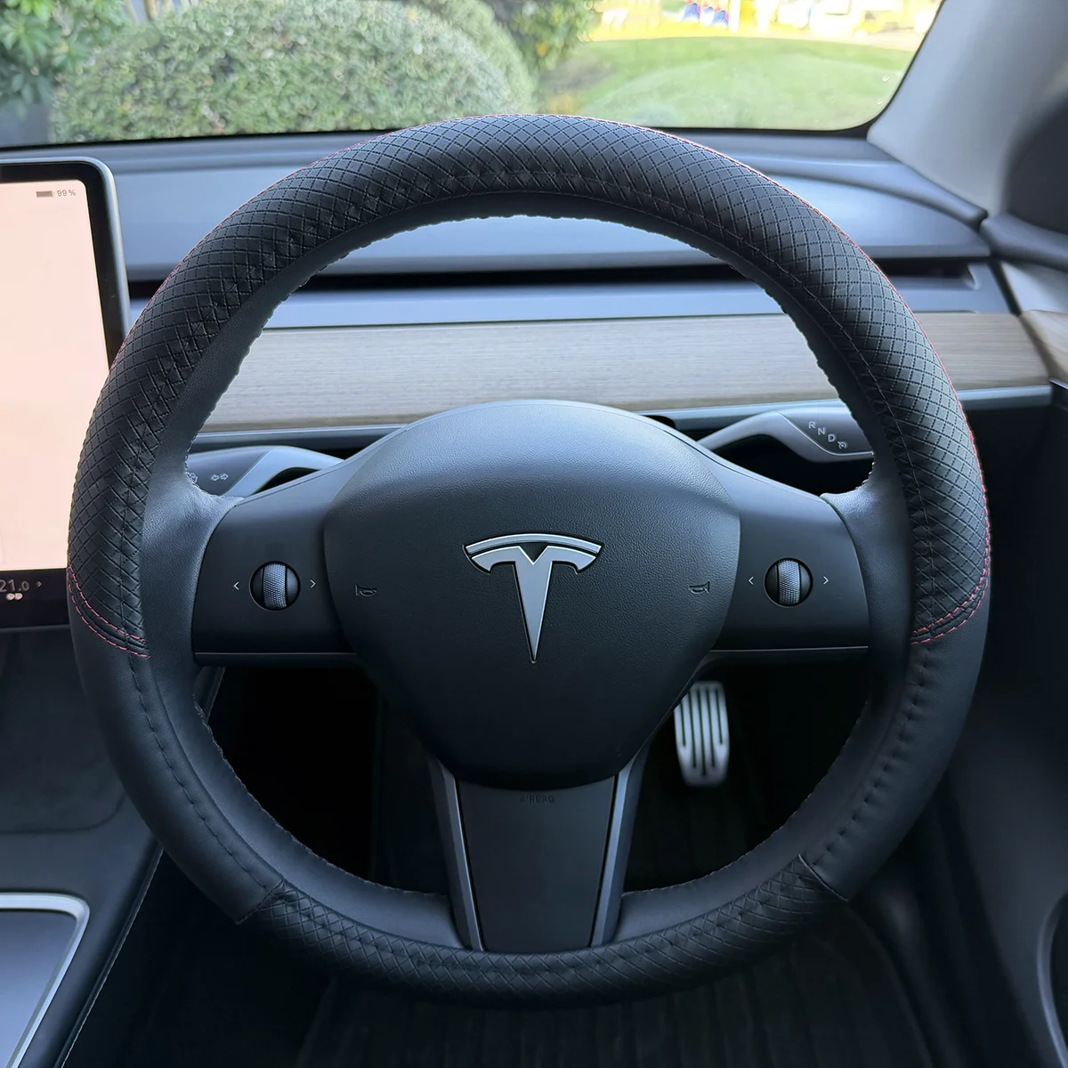 Tesla steering wheel with a black diamond-patterned cover inside a car, with a wood-grain dashboard and large touchscreen visible to the left