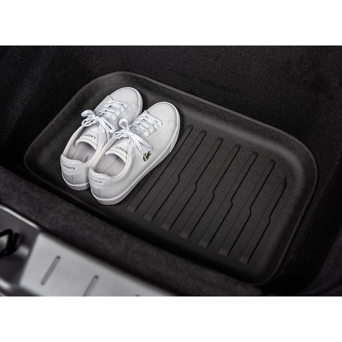 White Lacoste sneakers resting on a black car trunk mat inside the vehicle, with the crocodile logo visible on the side.