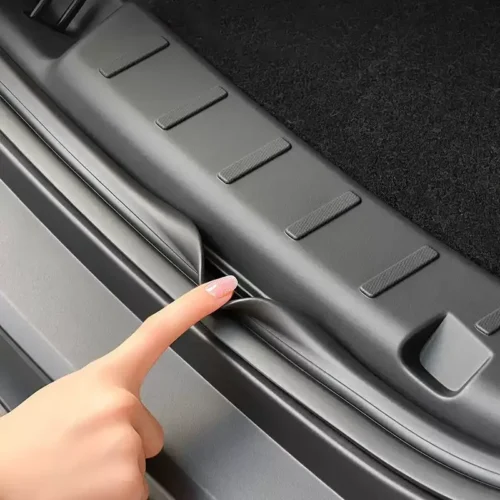 Close-up of a finger lifting the car's trunk sill cover along the cargo area edge, showing the textured step pads and rubber seal nearby.