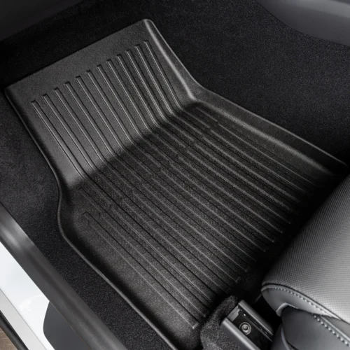 Black rubber car floor mat with ridged grooves in a driver’s footwell, resting over dark carpet near the seat edge.