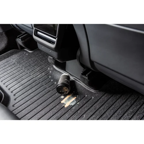 Camera lens lying on a ribbed car floor mat near the center console, with a coffee spill nearby.