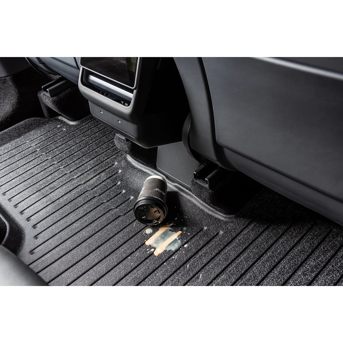 Camera lens lying on a ribbed car floor mat near the center console, with a coffee spill nearby.