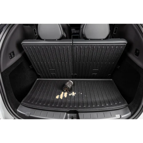 Open car trunk with folded rear seats and ribbed cargo mat; a coffee spill and a camera lens sit on the mat, showing clutter in storage space.