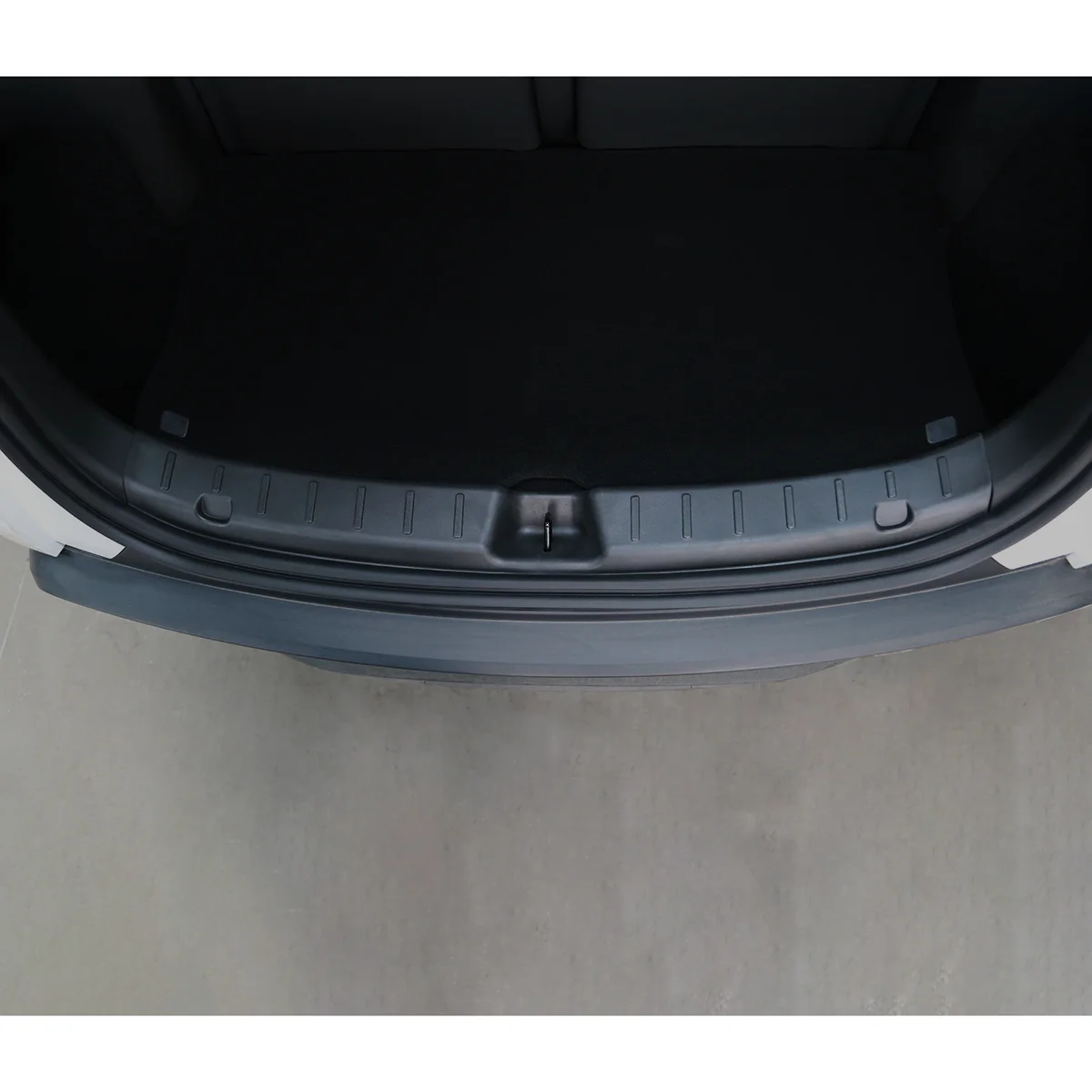 Open car trunk seen from above, showing a dark empty cargo area and a ridged plastic scuff plate along the edge with a central latch