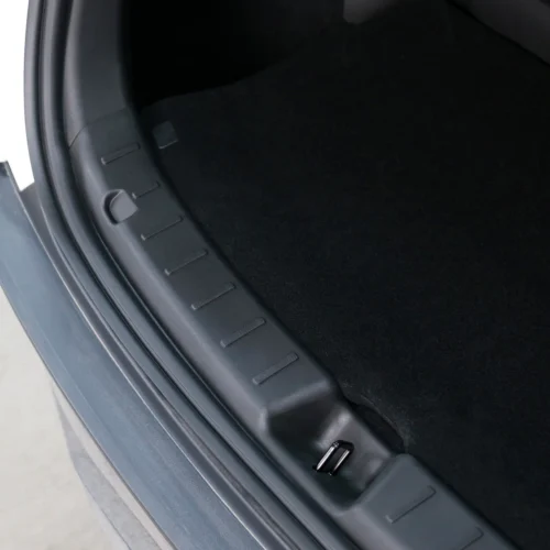 Close-up of a car trunk opening showing a black plastic scuff plate with grip recesses and a small metal latch at the edge of the carpeted trunk interior
