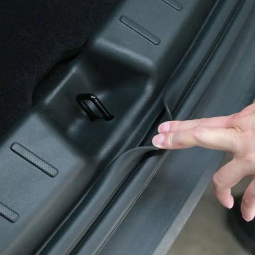 Close-up of a car trunk interior showing a small black metal hook attached to the frame; a person’s fingers pull back the rubber weather seal near the hinge area.